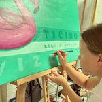 Load image into Gallery viewer, Person painting on a large green canvas with text and a flamingo design.
